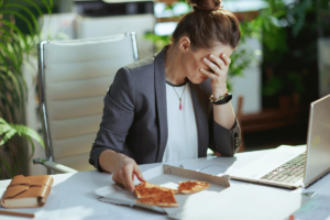 distraught looking professional woman seated at her laptop eating cold, leftover pizza for lunch and pondering how depression can affect your diet.