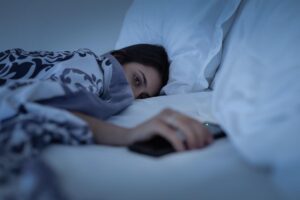 woman who is unaware of the connection between childhood trauma and sleep issues lying awake in her bed with cell phone clutched protectively in her hand