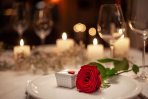 A red rose is laying across a white plate on a beautifully decorated table top with candlelight in the background. There is a small gift next to the rose, wrapped in white and gold. There are no people at the table.