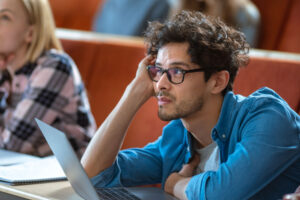 A male student is sitting in his college classroom with his laptop open. He's having difficulty concentrating due to ADHD burnout.