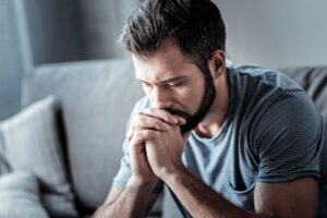 A man is sitting with his hands folded against his chin. He is experiencing signs of chronic depression.