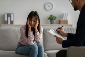 A woman is sitting on a couch and has her hands pressed to her temples. She is sitting across from a therapist.