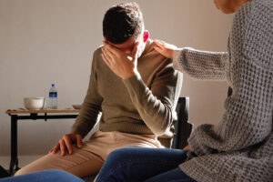 A man is sitting in a chair with his hand to his face. He seems to be crying. Another person has their hand on his shoulder, offering him comfort.